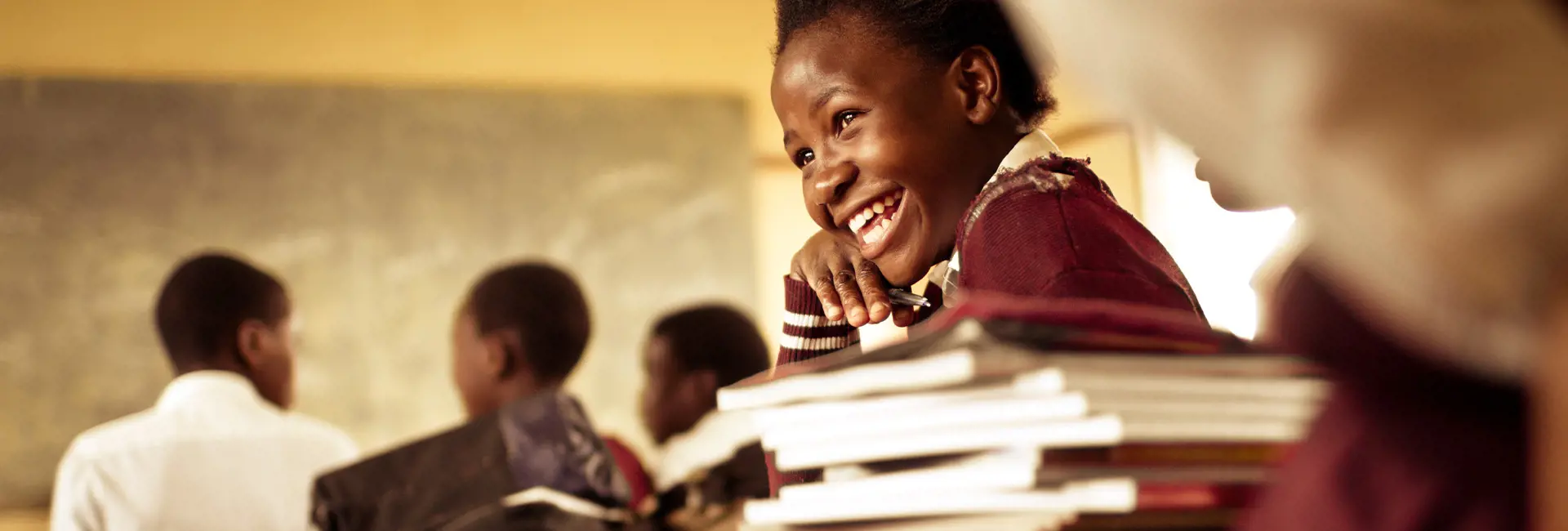Students in a classroom setting with a focus on a student in the foreground wearing a maroon sweater and holding a stack of books