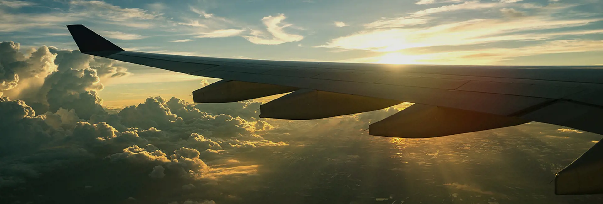 View of an airplane wing during flight at sunset, with golden sunlight illuminating clouds and the landscape below.