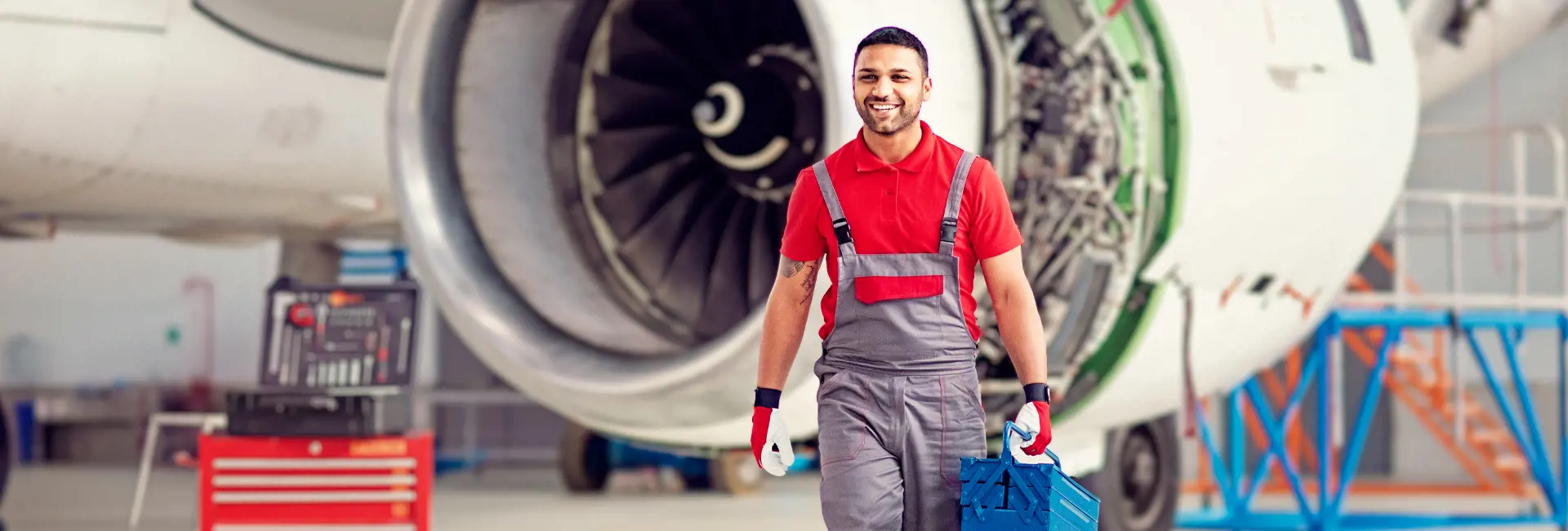Smiling aviation technician walking with a toolbox in front of an airplane engine in a hangar