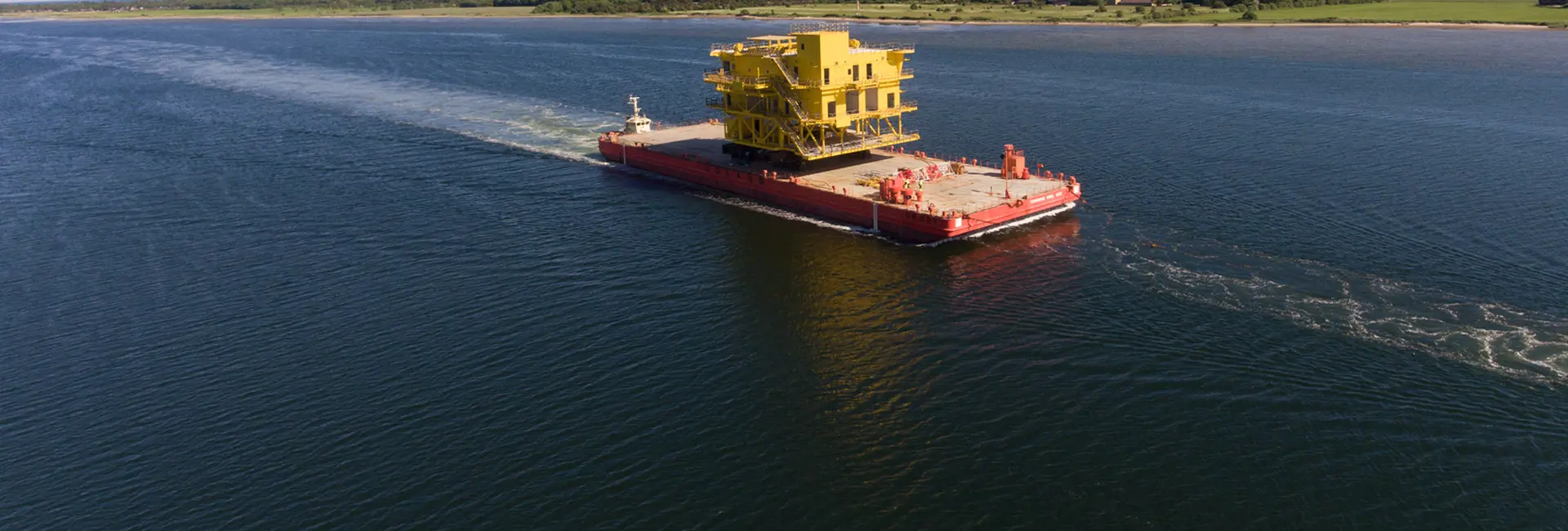 A large barge transporting a massive yellow industrial structure across a body of water, with greenery in the background.