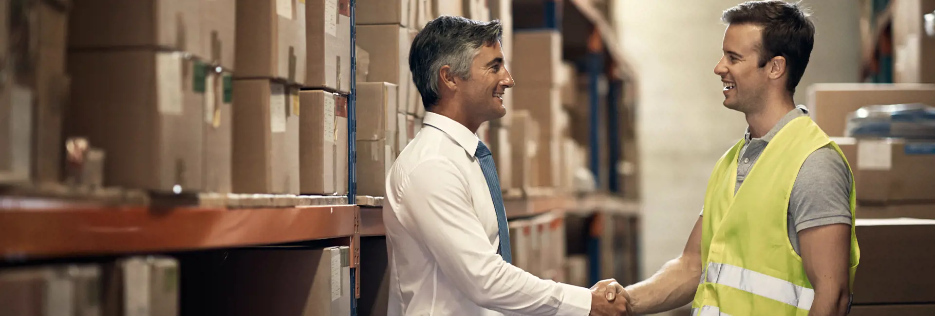 Two professionals shaking hands in a warehouse with stacked boxes