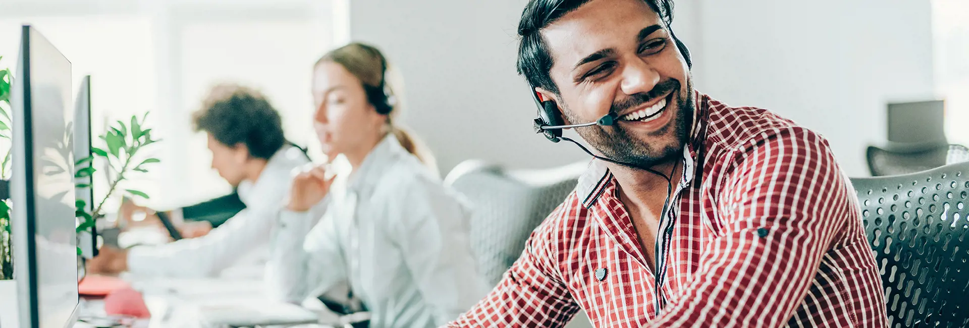 Smiling customer service representative wearing a headset in an office with colleagues working in the background