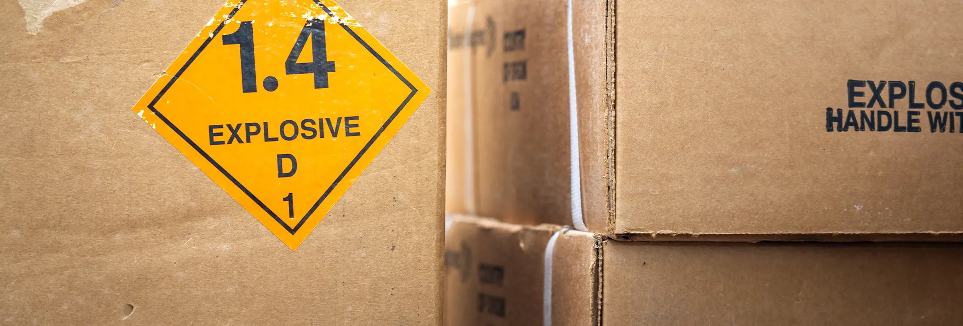 Cardboard boxes labeled with hazardous material warnings, including a yellow '1.4 Explosive' sign