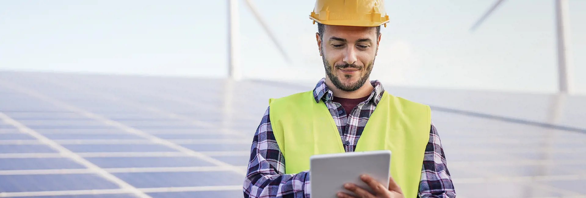 Engineer in a hard hat and safety vest using a tablet near solar panels and wind turbines