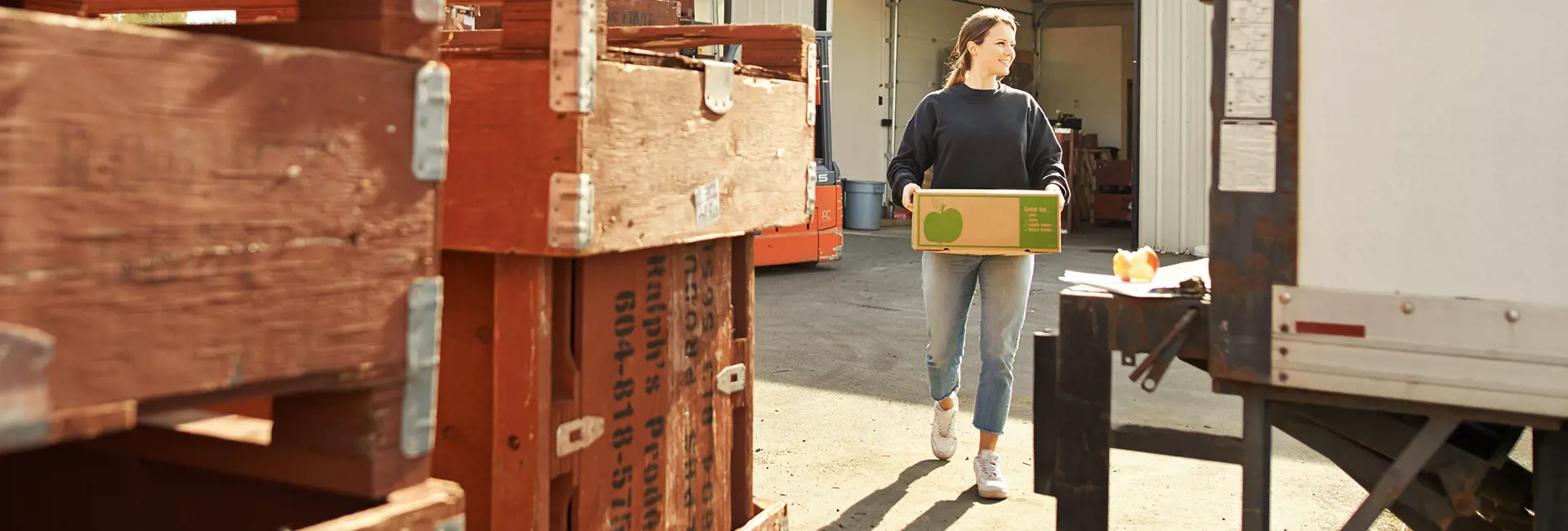 Smiling woman carrying a produce box near wooden crates and a loading dock