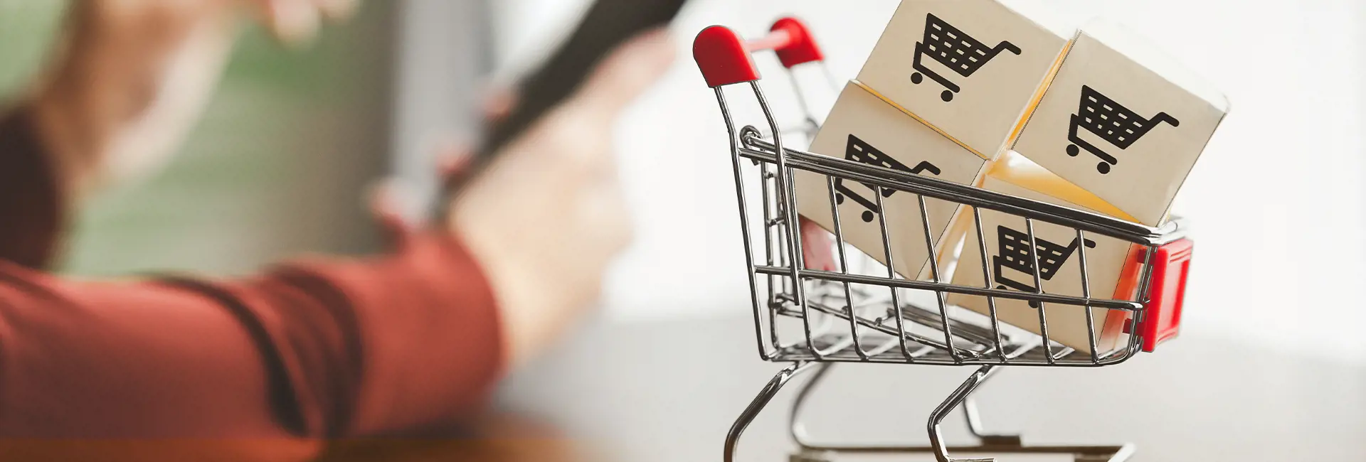 "A miniature shopping cart filled with small cardboard boxes, each featuring a shopping cart icon, with a blurred figure holding a tablet or smartphone in the background