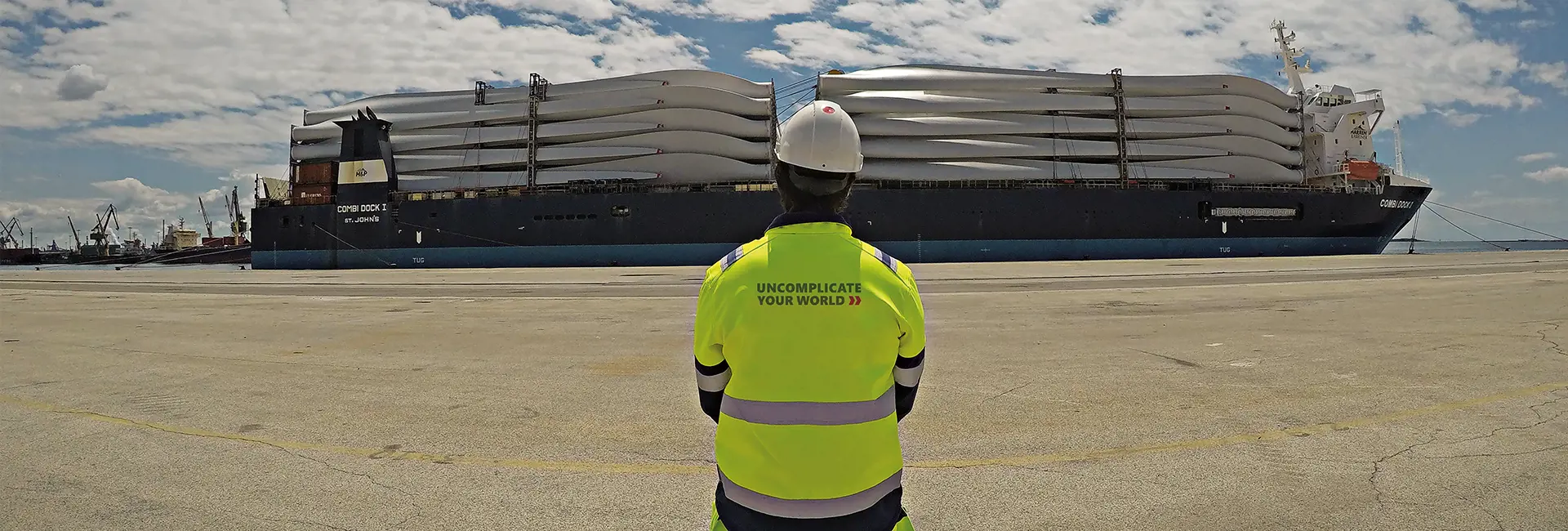 Logistics professional in a high-visibility jacket overseeing a cargo ship loaded with wind turbine blades at a port