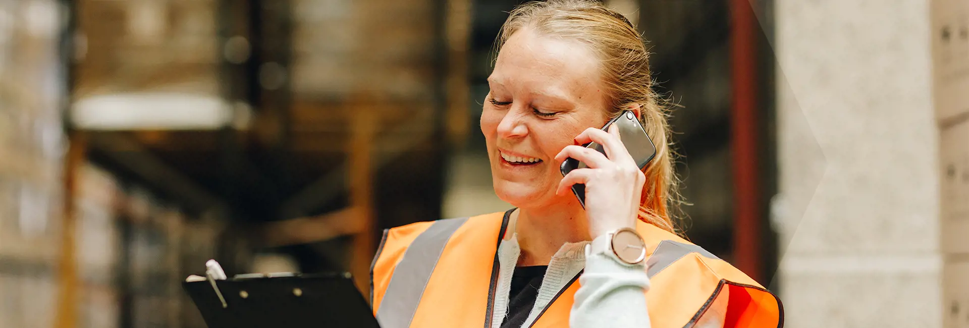 A scan global logistics worker wearing an orange high-visibility vest holds a clipboard and talks on a mobile phone in a warehouse.