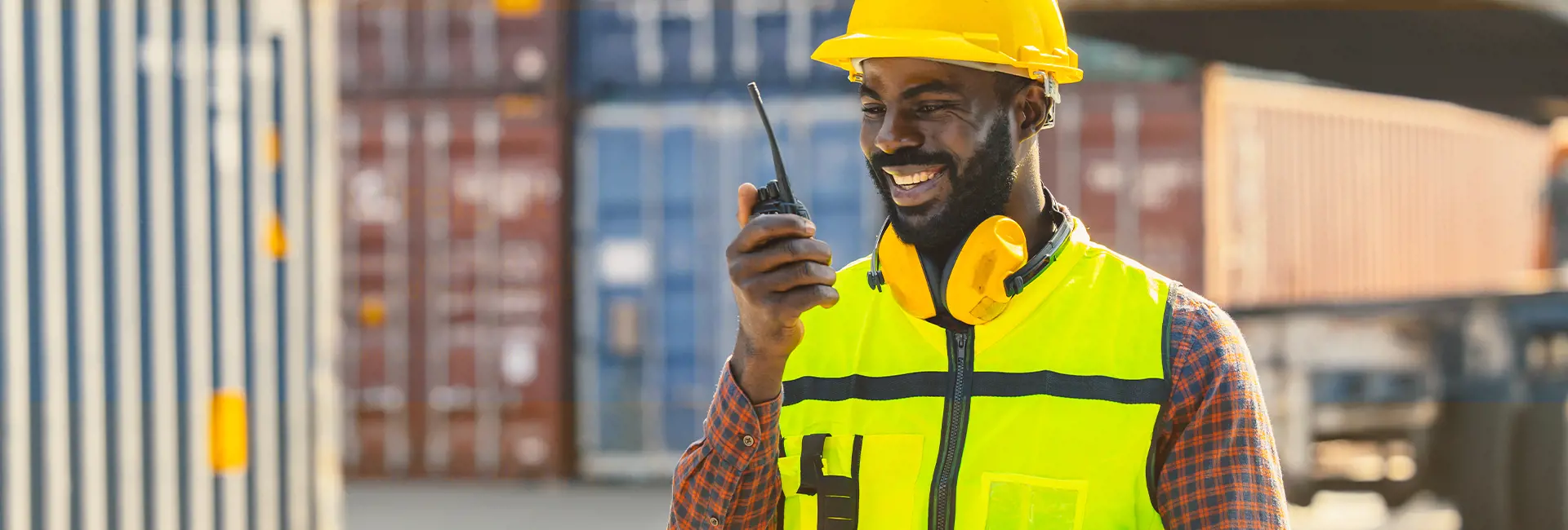 A logistics professional in a yellow safety vest, hard hat, and protective headphones is smiling while using a walkie-talkie at a port. Stacked shipping containers and part of a truck are visible in the background, showcasing efficient communication and coordination in ocean freight operations.