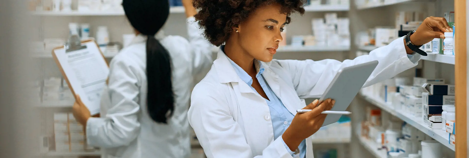 Pharmacist organizing medicine on shelves while using a tablet, representing Scan Global Logistics' pharma healthcare logistics