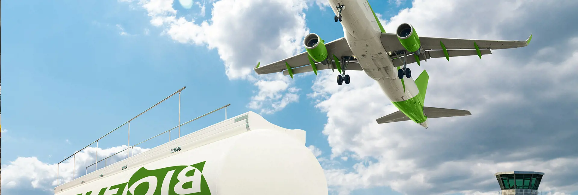 An airplane with a green and white color scheme flying over a large white fuel tank labeled with green text, under a partly cloudy sky