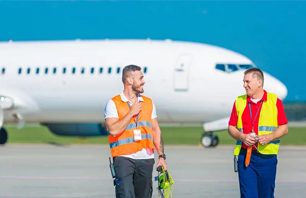 Two airfreight professionals in high-visibility vests walking on an airport tarmac, with an airplane in the background