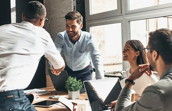 Business professionals in a meeting, two shaking hands across a table with laptops