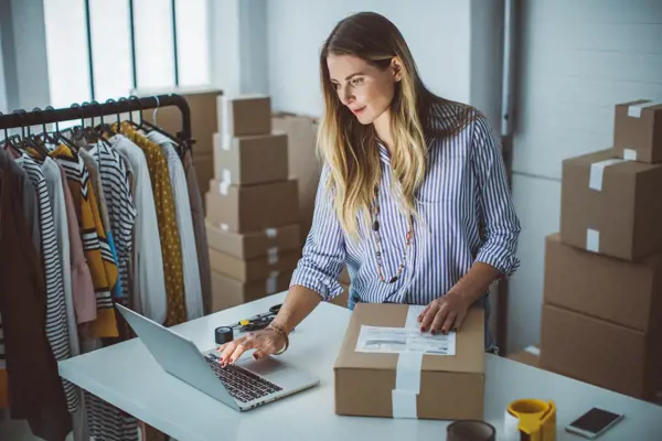 Businesswoman in a warehouse using a laptop to manage shipments, with packages and clothing racks in the background