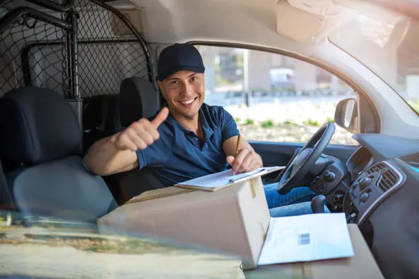 Smiling delivery driver giving a thumbs-up from a van, with packages and a clipboard in the front seat
