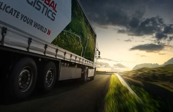 A large logistics truck driving on a road at sunset, labeled 'SCAN GLOBAL LOGISTICS' with a green landscape and winding roads on its side.