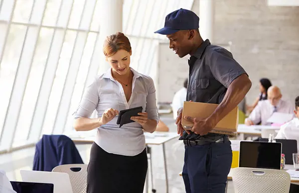 An onboard courier delivering a package to a businesswoman in an office, with the two reviewing a tablet.