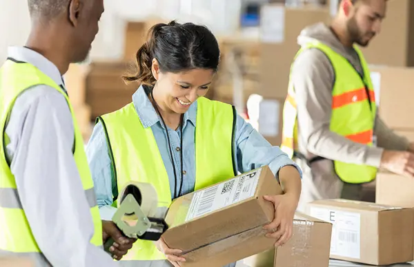 Three workers in high-visibility vests engaged in packaging activities in a warehouse, illustrating the picking, packing, and preparing of goods for delivery