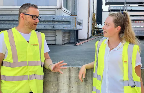 Two scan global logistics workers wearing yellow safety vests having a casual conversation near an industrial loading area