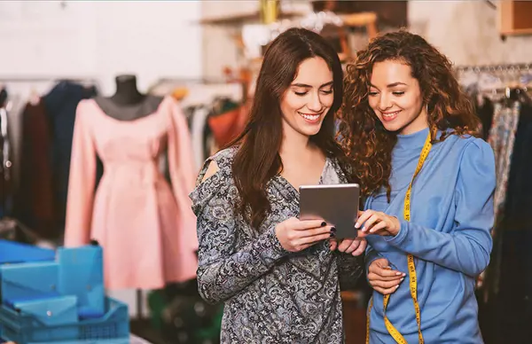 Two women in a clothing store reviewing information on a tablet, surrounded by garments and tailoring tools