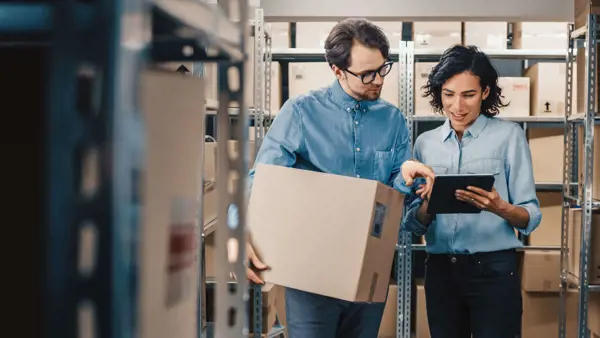 Two warehouse workers discussing inventory while reviewing a tablet, surrounded by shelves of boxes